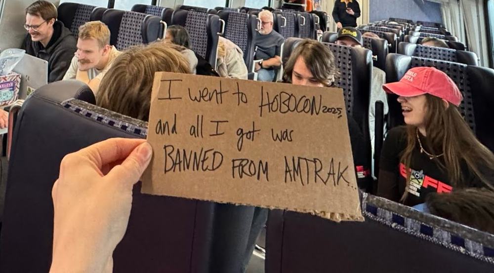A hand-lettered cardboard sign reading "I went to HOBOCON 2025 and all I got was BANNED FROM AMTRAK" held up in the train car