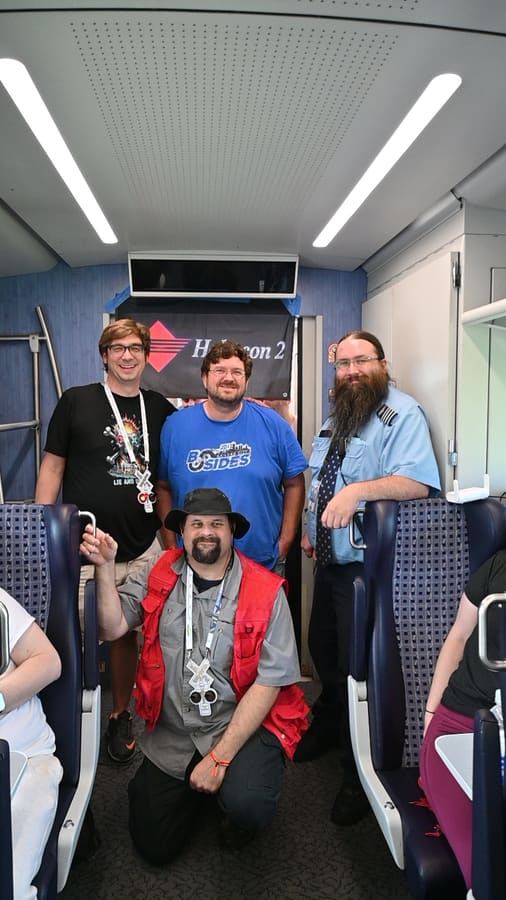 Four attendees posing in the aisle of the train car beneath a Hobocon 2 banner taped to the bulkhead, one kneeling in a red vest, another in a BSides Kansas City shirt