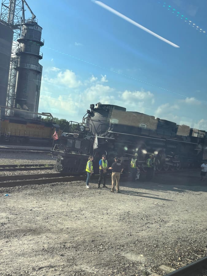 Union Pacific Big Boy steam locomotive number 4014 stopped on a siding beside grain silos, with crew in hi-vis vests standing around it, photographed through a moving train window